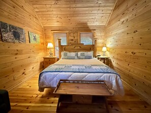 Un dormitorio de cabaña de madera con una cama, dos mesitas de noche, una lámpara y un banco.