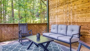 Un espacio de estar al aire libre techado, con pared de madera, un banco, una mesa y vistas a un bosque.