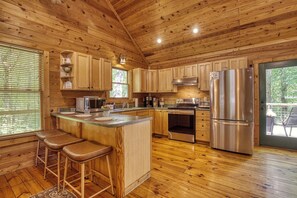 Interior de una cabaña de madera con cocina, que incluye un refrigerador de acero inoxidable, una estufa y un lavamanos. Se observan armarios de madera y una zona con taburete de bar.