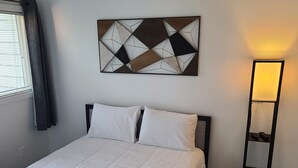 Una cama bien hecha con sábanas blancas, un cuadro abstracto en la pared sobre ella, y una mesita de noche con una lámpara.