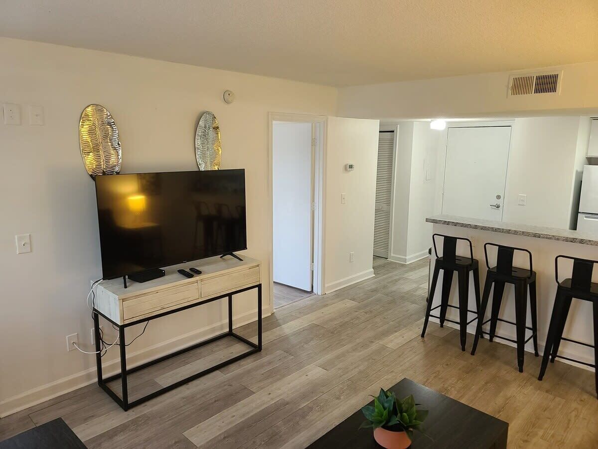 A modern living room with a flat-screen TV on a white console, a coffee table with a plant, and a kitchen area with a bar stool and a refrigerator.