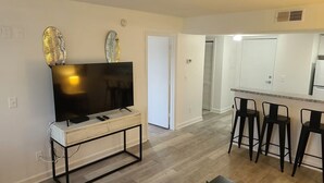 Una sala de estar moderna con un televisor de pantalla plana sobre una consola blanca, una mesa de centro con una planta, y una zona de cocina con un taburete alto y un refrigerador.