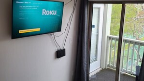 Una habitación con un televisor montado que muestra instrucciones para activar Roku TV, un balcón con vistas a zonas verdes y una cama con colchón blanco.