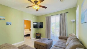 A living room with a ceiling fan, a flat-screen TV, and a large window with curtains.
