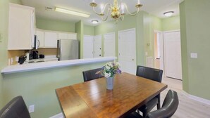 A dining area with a wooden table, chairs, a chandelier, and a kitchen in the background.