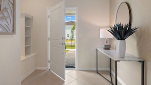 Een hal met een witte deur die uitkomt op een uitzicht op een huis, een console tafel met een lamp en een plant, en een spiegel aan de muur.