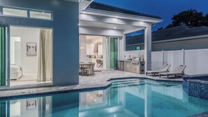 Une maison moderne dotée d’une piscine, d’un patio avec des chaises longues et d’un espace cuisine visible à travers des portes vitrées.