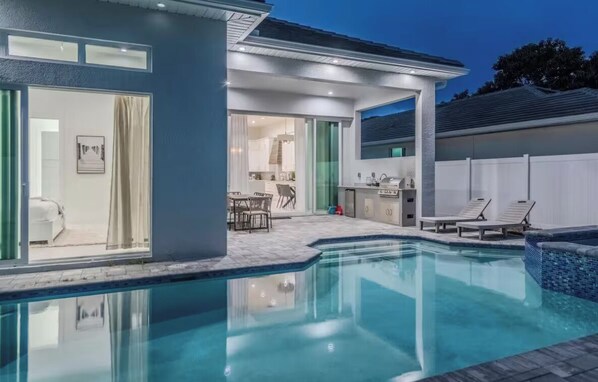 A modern house with a swimming pool, a patio with lounge chairs, and a kitchen area visible through glass doors.