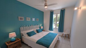 A bedroom with teal walls, a white bed with blue bedding, a ceiling fan, and a window with curtains. - Orient blue garden, direct access 50m from the beach! (Baie orientale)