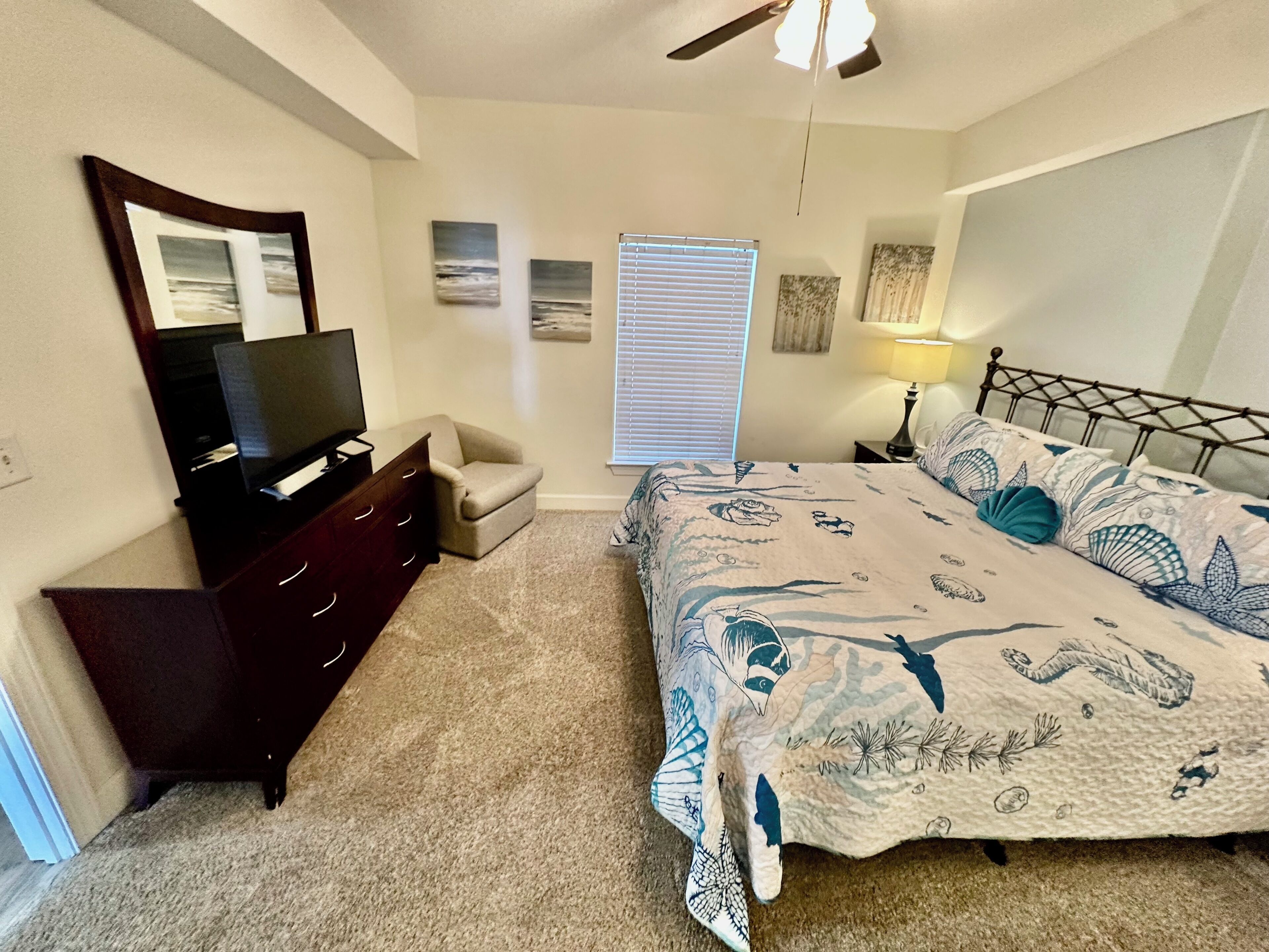 A bedroom with a bed, a dresser, a TV, a chair, a ceiling fan, and artwork on the walls.