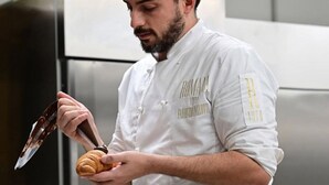 Un chef con uniforme blanco y la inscripción "ROMA 1970 FABBRIO RALLOTTI" en la manga, prepara pasteles en una cocina con un refrigerador de acero inoxidable al fondo.