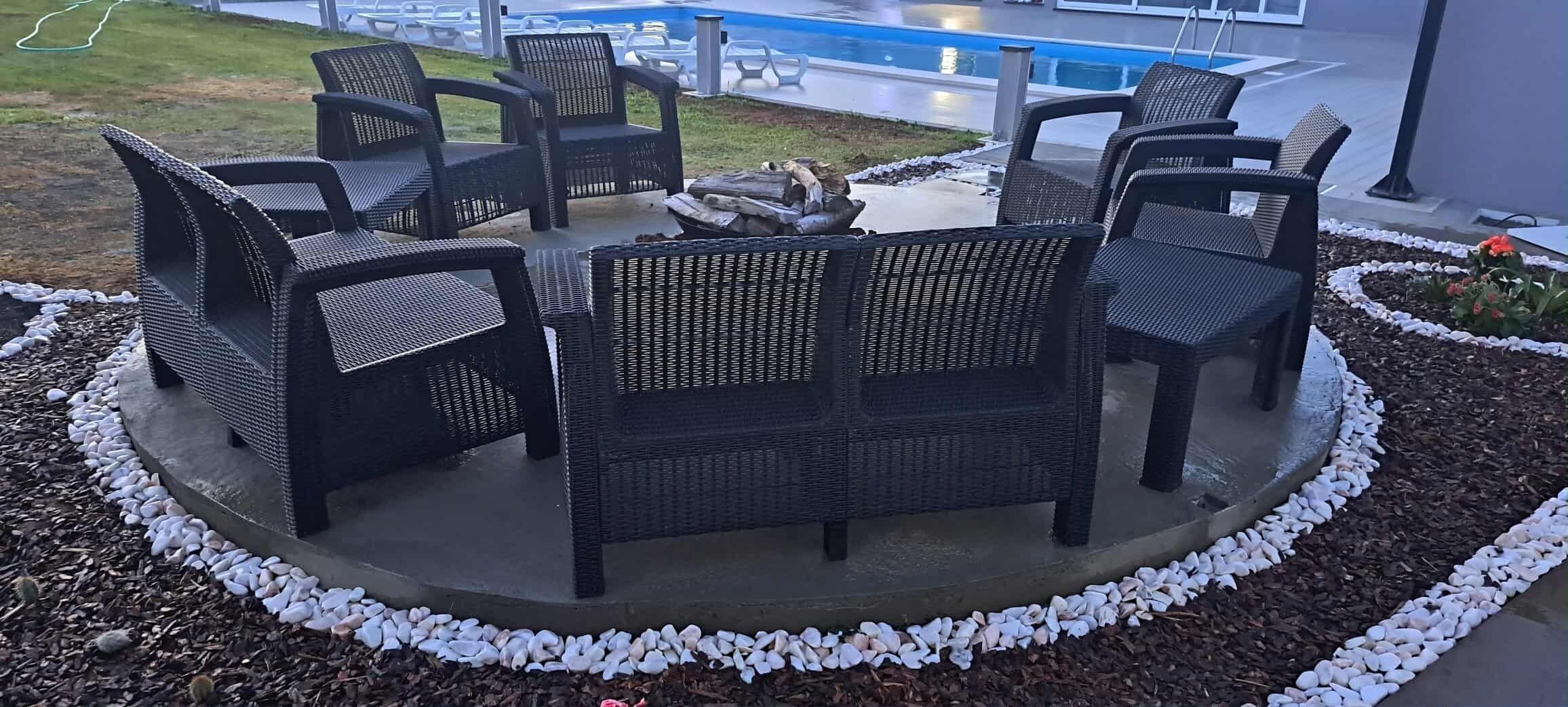 A circular outdoor seating area with wicker chairs and a central fire feature, surrounded by white pebbles and mulch.