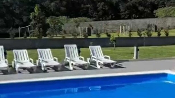 Una piscina con sillas reclinables blancas y un área verde con árboles al fondo.