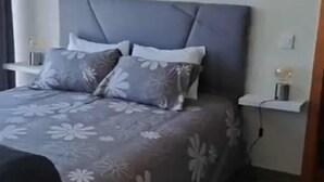 Una cama bien hecha con un cabecero gris, dos almohadas y una colcha estampada con motivos florales. Hay una mesita de noche con una lámpara y un enchufe en la pared.