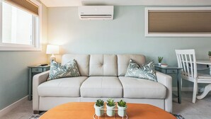 A living room with a beige sofa, a coffee table with potted plants, a side table with a lamp, and a wall-mounted air conditioner.