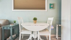 A dining area with a white table and chairs, a small side table with a plant, and a window with blinds.