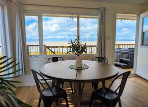 Une table de salle à manger ronde, entourée de quatre chaises, agrémentée d’une plante en pot, et offrant une vue sur la plage grâce à de grandes fenêtres.