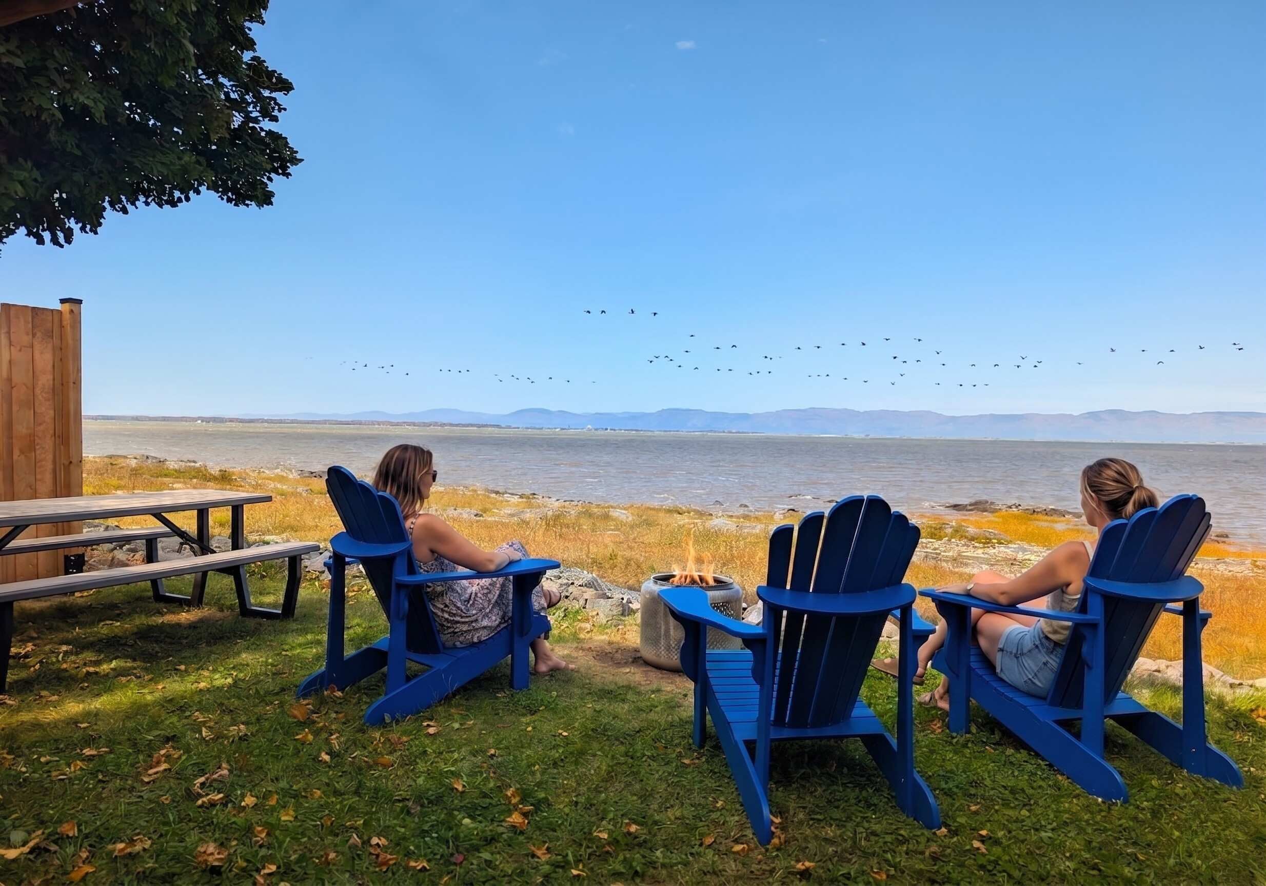 Two Adirondack chairs facing a lake with a picnic table and a fire pit nearby.