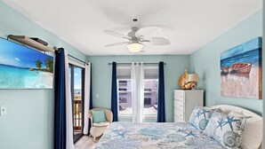 A bedroom with a bed, a ceiling fan, a window with curtains, a chair, and a painting on the wall. - Spacious condo, across the street from the beach + Free Attraction Tickets! (North Myrtle Beach)