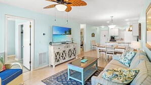 A spacious living room with a ceiling fan, a television, a dining area, and a kitchen in the background. - Spacious condo, across the street from the beach + Free Attraction Tickets! (North Myrtle Beach)