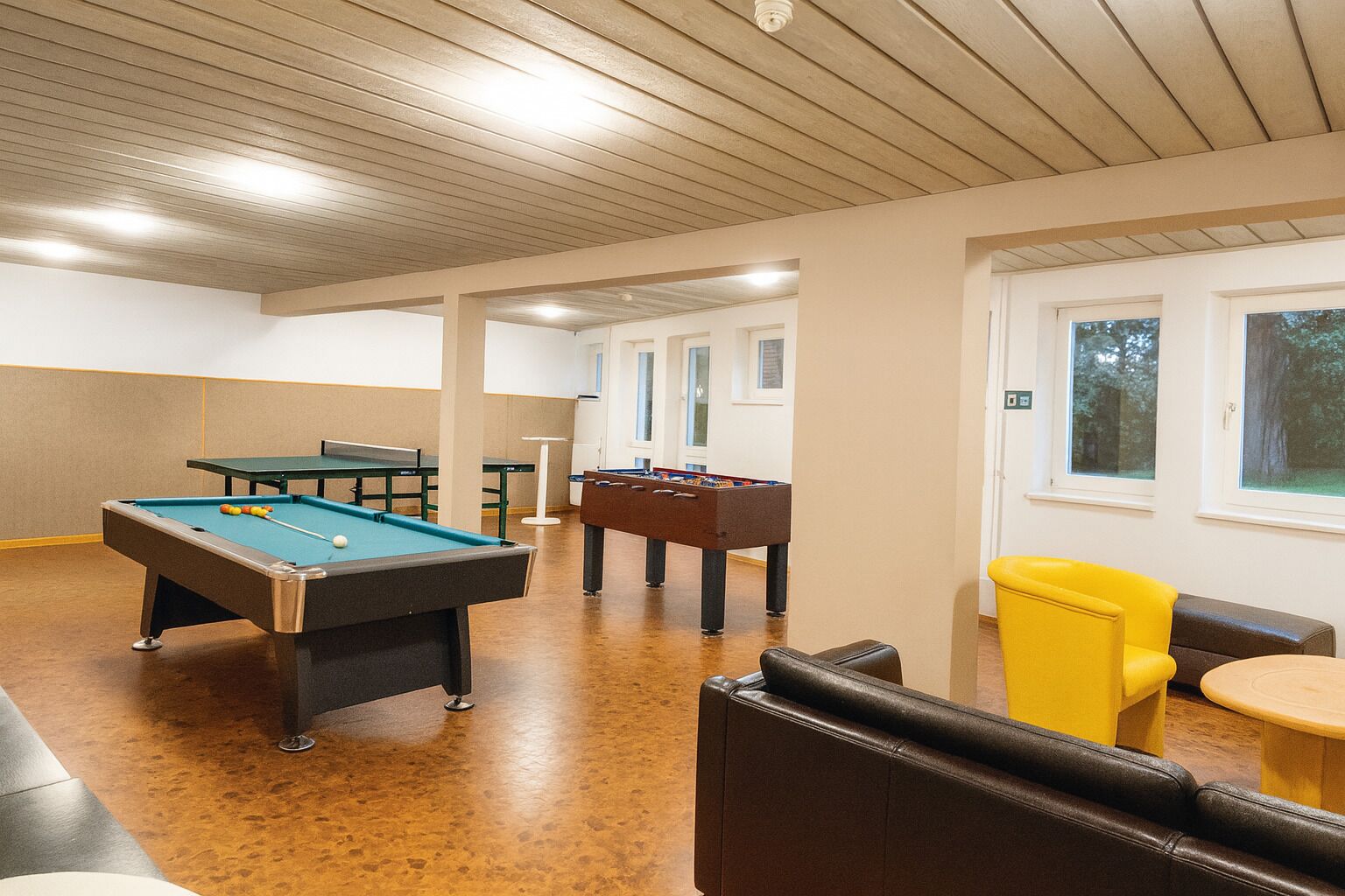 Mesa de billar, mesa de fútbol de mesa y zona de estar en un espacio interior bien iluminado con ventanas.