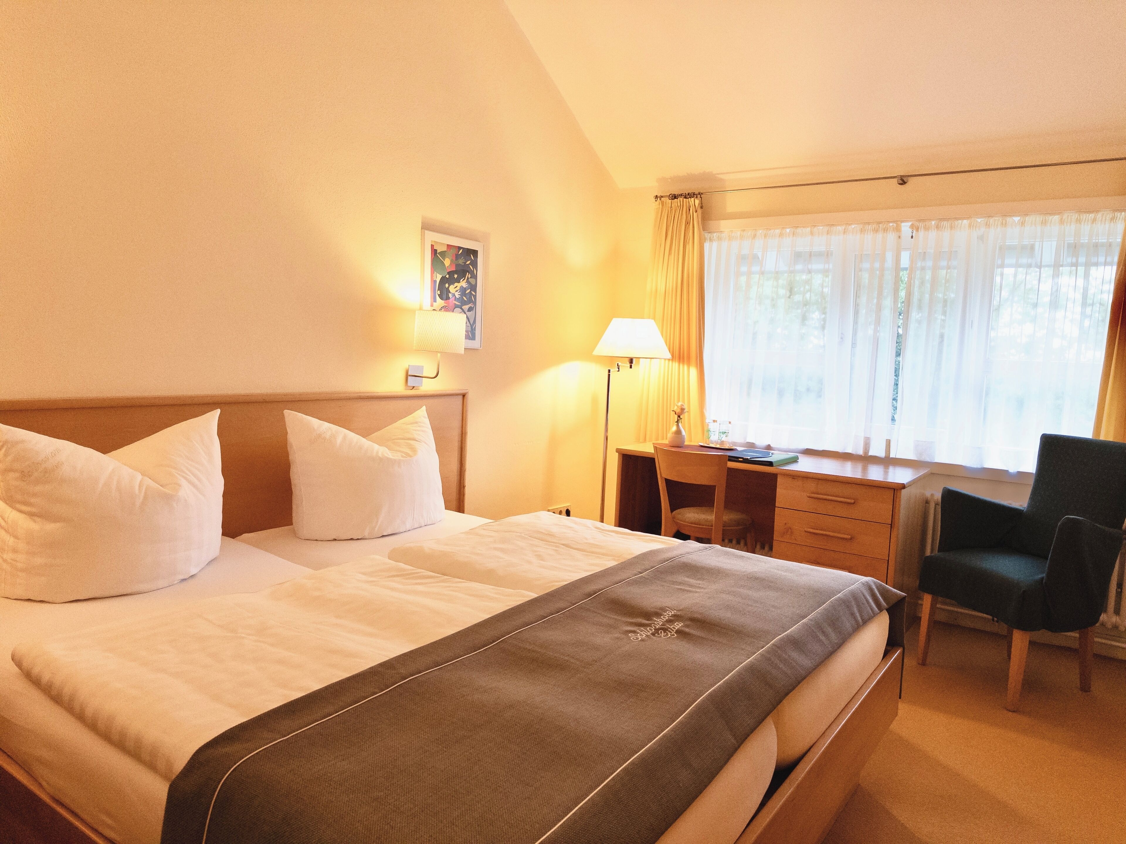 A hotel room with a bed, a desk, a chair, a lamp, and a window with curtains.