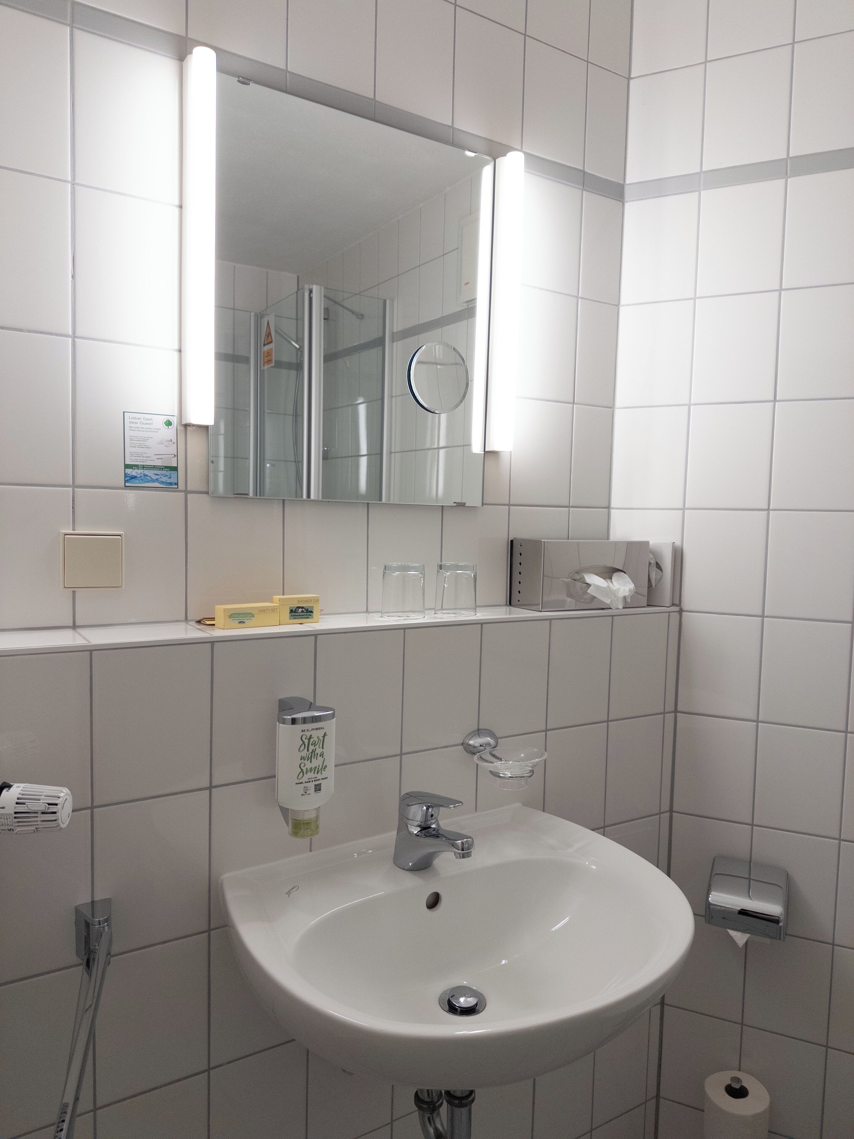 A bathroom with a white sink, mirror, and tiled walls. There is a soap dispenser, a glass soap dish, and a toilet paper holder.