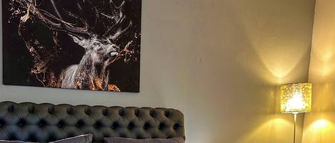A bedroom with a tufted headboard, multiple pillows, a bed with a grey and blue bedspread, a wall-mounted painting of a deer, and a floor lamp.