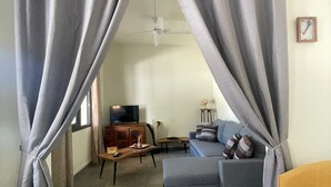 Una sala de estar con cortinas grises, un sofá, un sillón amarillo, un televisor y un ventilador de techo.