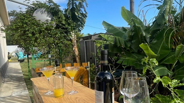 Una mesa de madera con una botella de vino y copas, colocada al aire libre con vegetación y un cielo despejado.