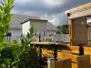 Een houten terras met een tafel, een wit gebouw met een grijs dak, en een bamboestructuur op de achtergrond.