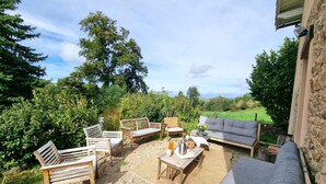 Un espace extérieur aménagé avec des meubles en bois, un canapé gris et une table sur laquelle se trouvent une bouilloire et des aliments.