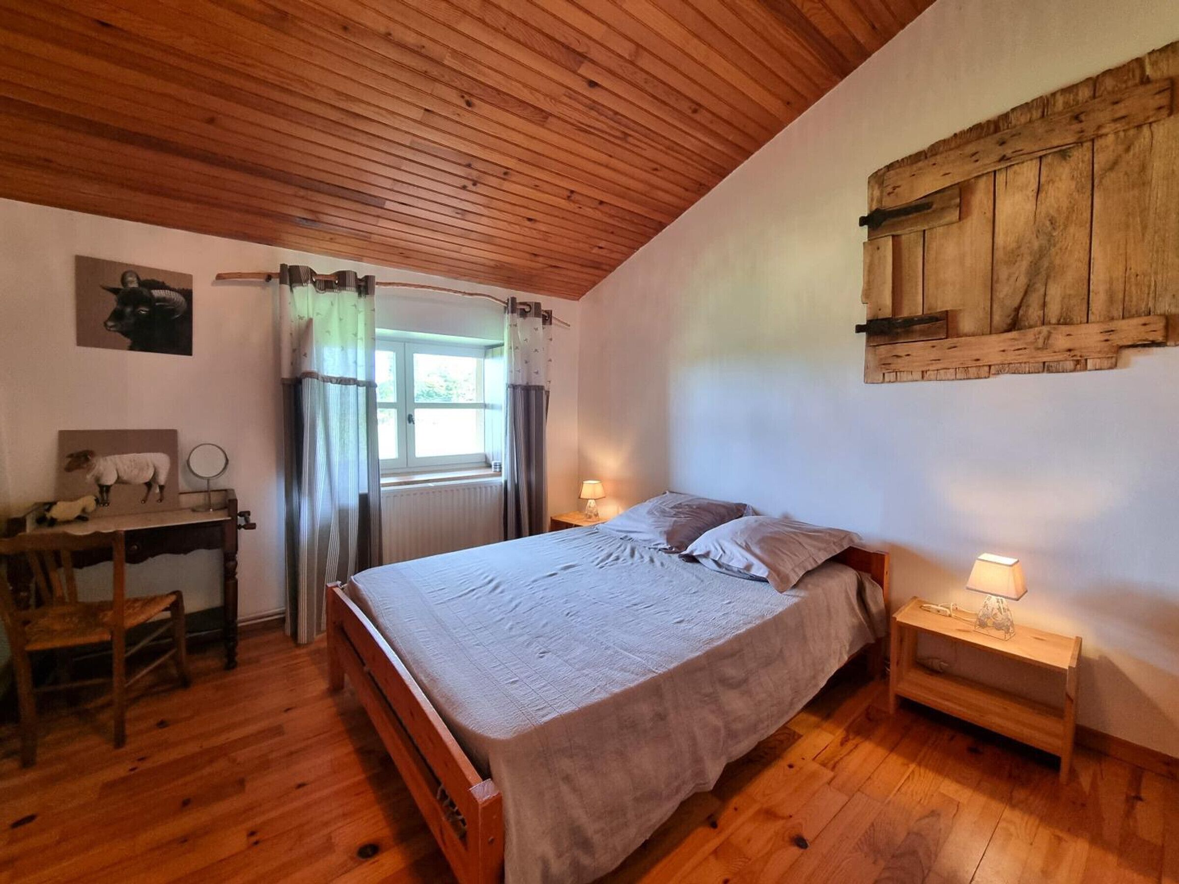 A cozy bedroom with a wooden ceiling, a bed with a grey bedspread, two bedside tables with lamps, a wooden desk with a chair, and a window with curtains.