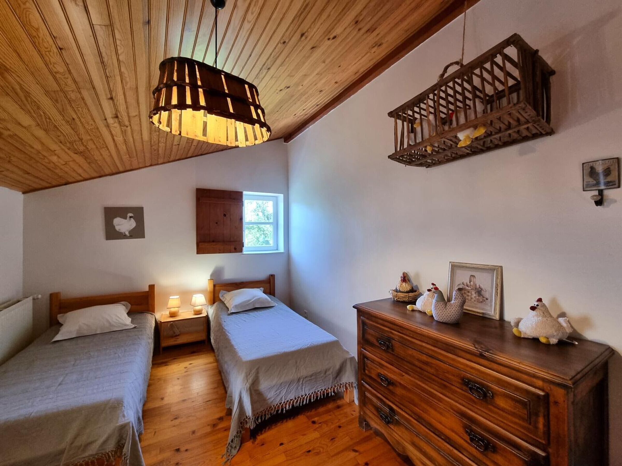 Une chambre confortable avec deux lits simples, une commode en bois, un luminaire suspendu et une petite fenêtre.