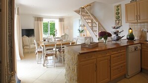 A kitchen with wooden cabinets, a dining area with white chairs, a staircase, and a view of greenery outside.