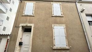 Un edificio con una puerta blanca, dos ventanas con contraventanas y un ventanal más pequeño debajo de las contraventanas.