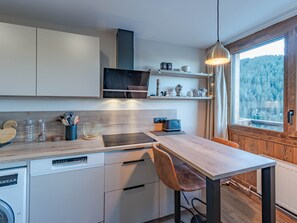 Una cocina moderna con lavavajillas, armarios de madera, una mesa de comedor con una silla y una ventana con vistas a un bosque.