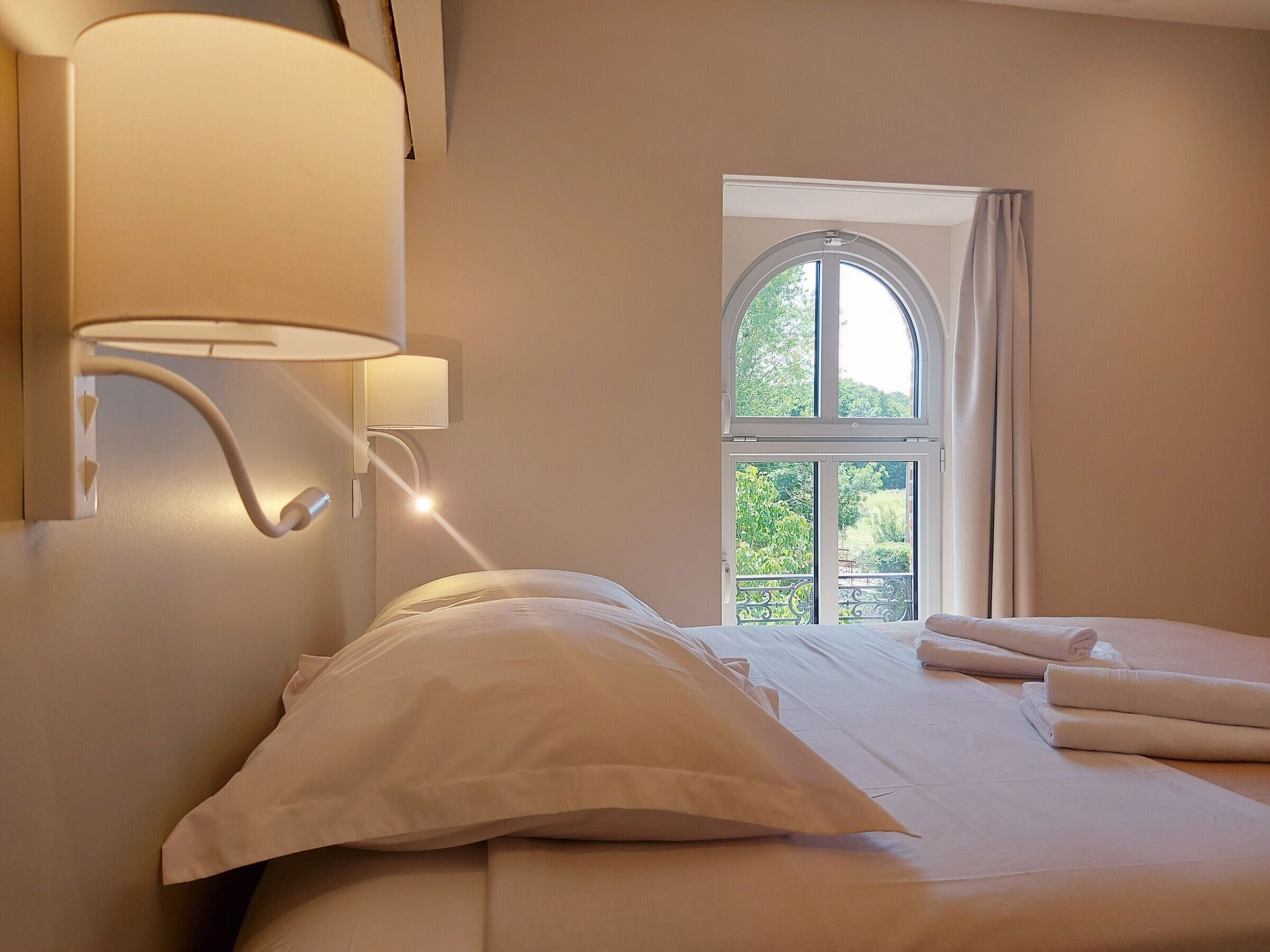 A hotel room with a bed, a wall-mounted lamp, and a window with a view of greenery.