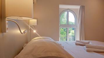 A hotel room with a bed, a wall-mounted lamp, and a window with a view of greenery.