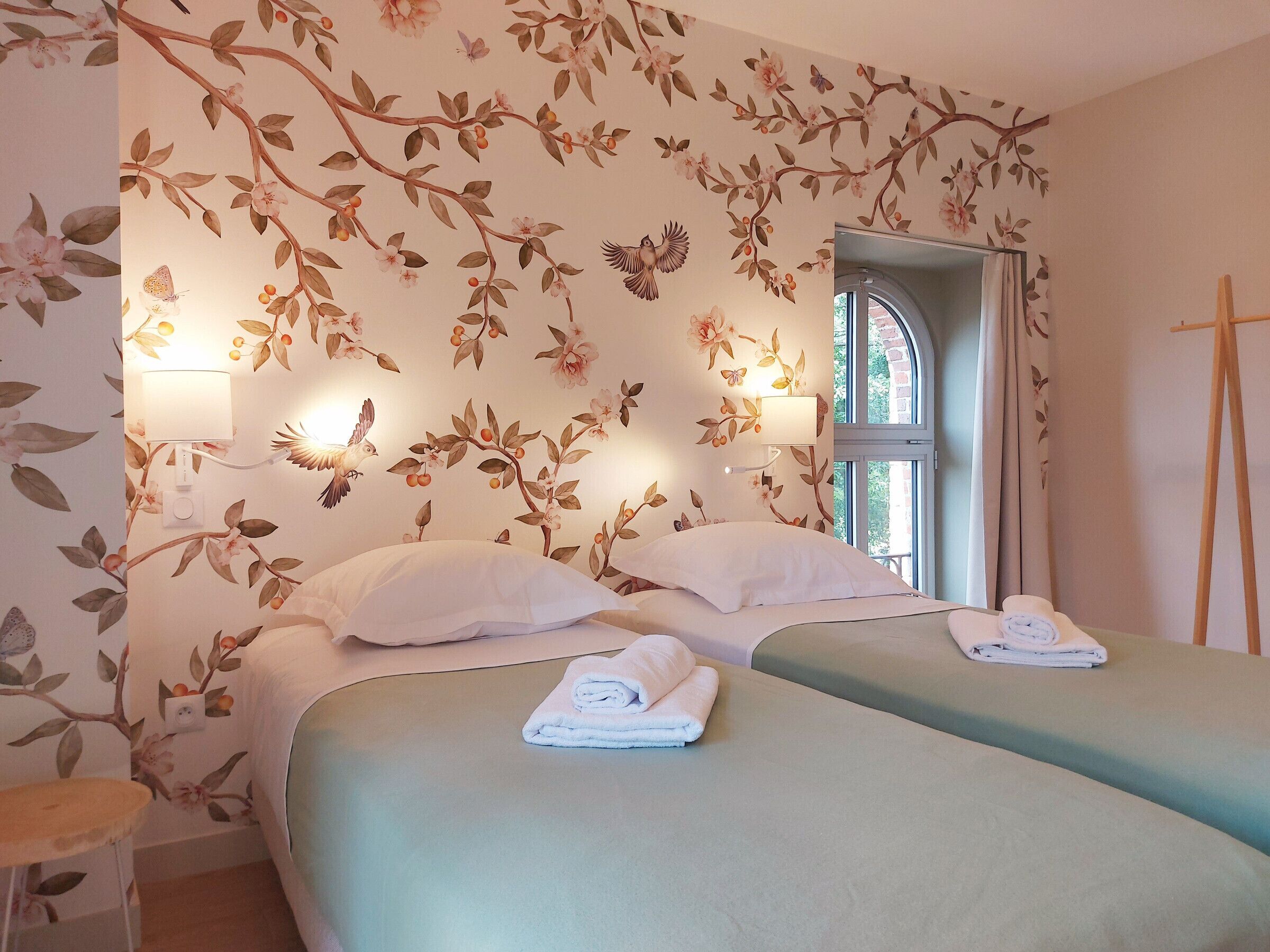 Habitació d'hotel amb dos llits, paper pintat floral, dues làmpades penjades a la paret, una finestra i un cavallet de fusta.
