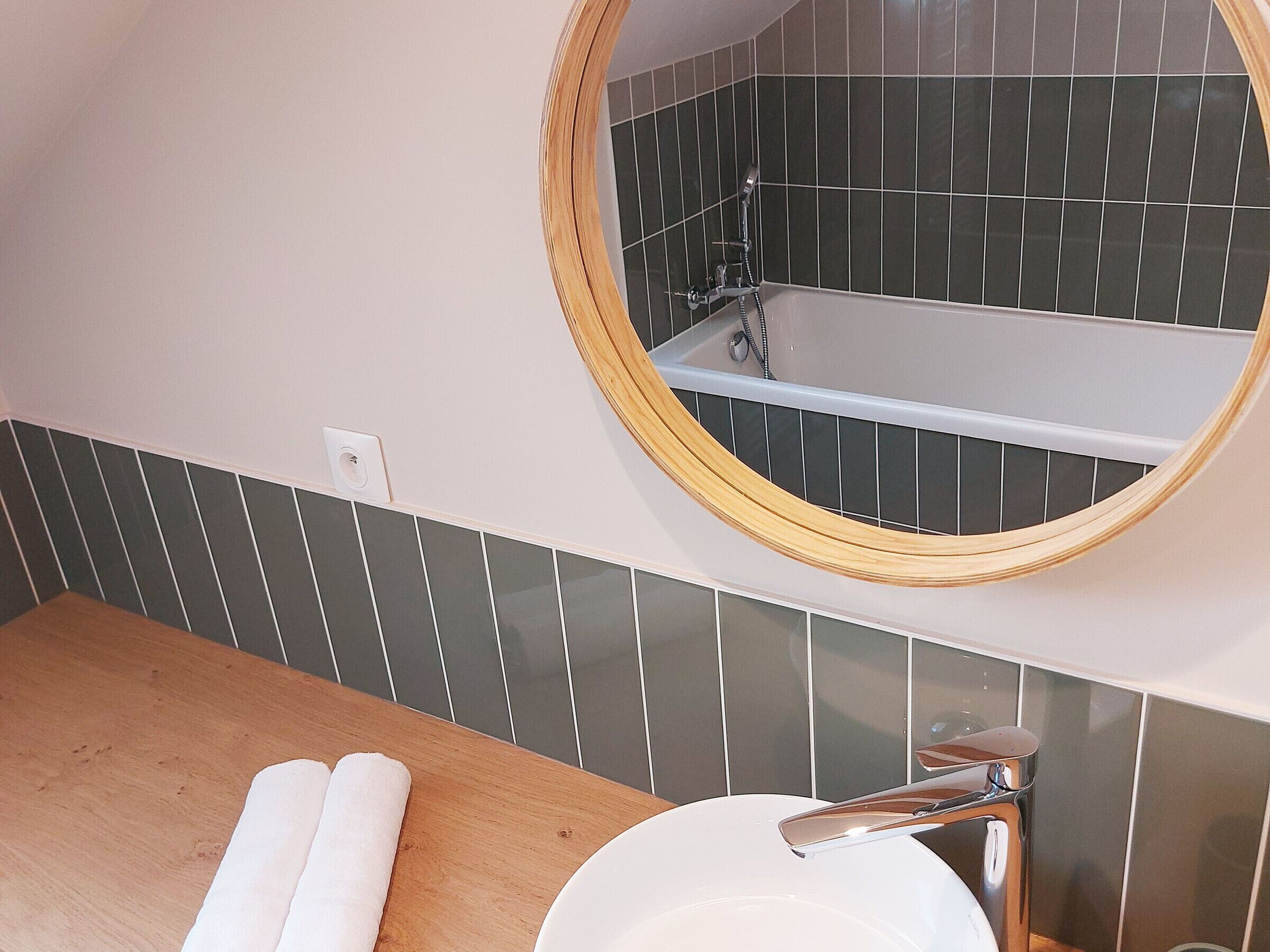 Une salle de bain avec un meuble-lavabo en bois, un miroir rond, un lavabo blanc et une baignoire équipée d’un pommeau de douche.