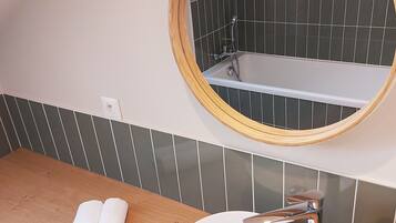 Une salle de bain avec un meuble-lavabo en bois, un miroir rond, un lavabo blanc et une baignoire équipée d’un pommeau de douche.