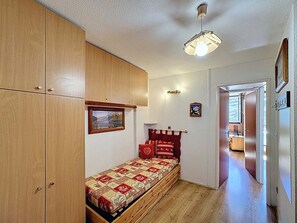 Quarto compacto com guarda-roupa de madeira, cama com colcha estampada, luminária pendente e quadro na parede.