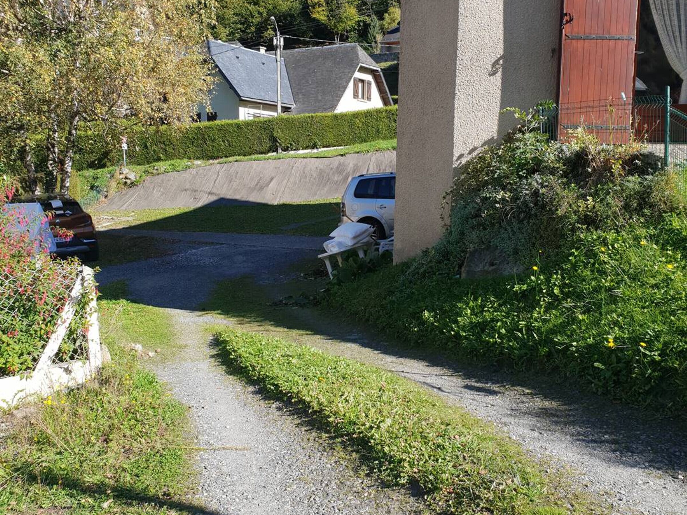 A gravel driveway leading to a parked car, a white building with a red door, and a house in the background.
