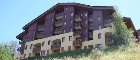 A multi-story building with balconies, likely a hotel or apartment complex, situated on a hillside with natural vegetation around.