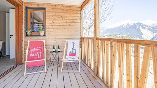A wooden deck with a view of snow-capped mountains, a small table with bottles, and a red banner. - Cozy chalet for 6 in nature (Aussois)