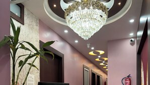 A reception area with a modern design, featuring a reception desk, a large chandelier, and a fire extinguisher mounted on the wall. - Hotel Anmol Inn (Karchana)