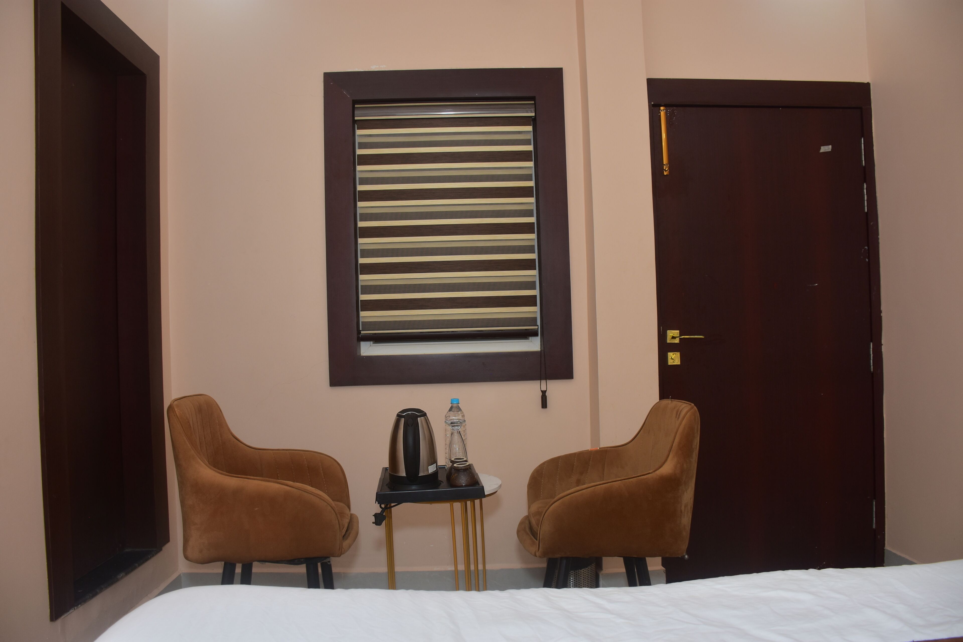 Una camera d'albergo con un letto, due poltrone marroni, un tavolino con un bollitore, una bottiglia e una finestra con persiane orizzontali.