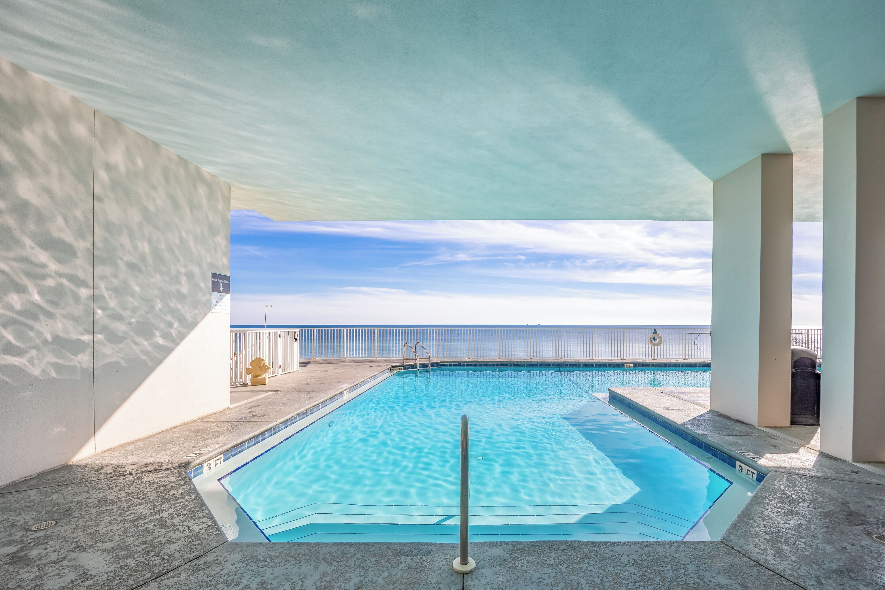 A swimming pool with clear blue water, surrounded by a white wall and a railing, with a view of the ocean in the background.