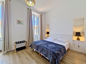 Une chambre à coucher avec un lit, une petite table, une chaise, une fenêtre avec des rideaux et une lampe fixée au mur.